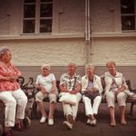 Five elderly women sitting on a park bench outside of a building.