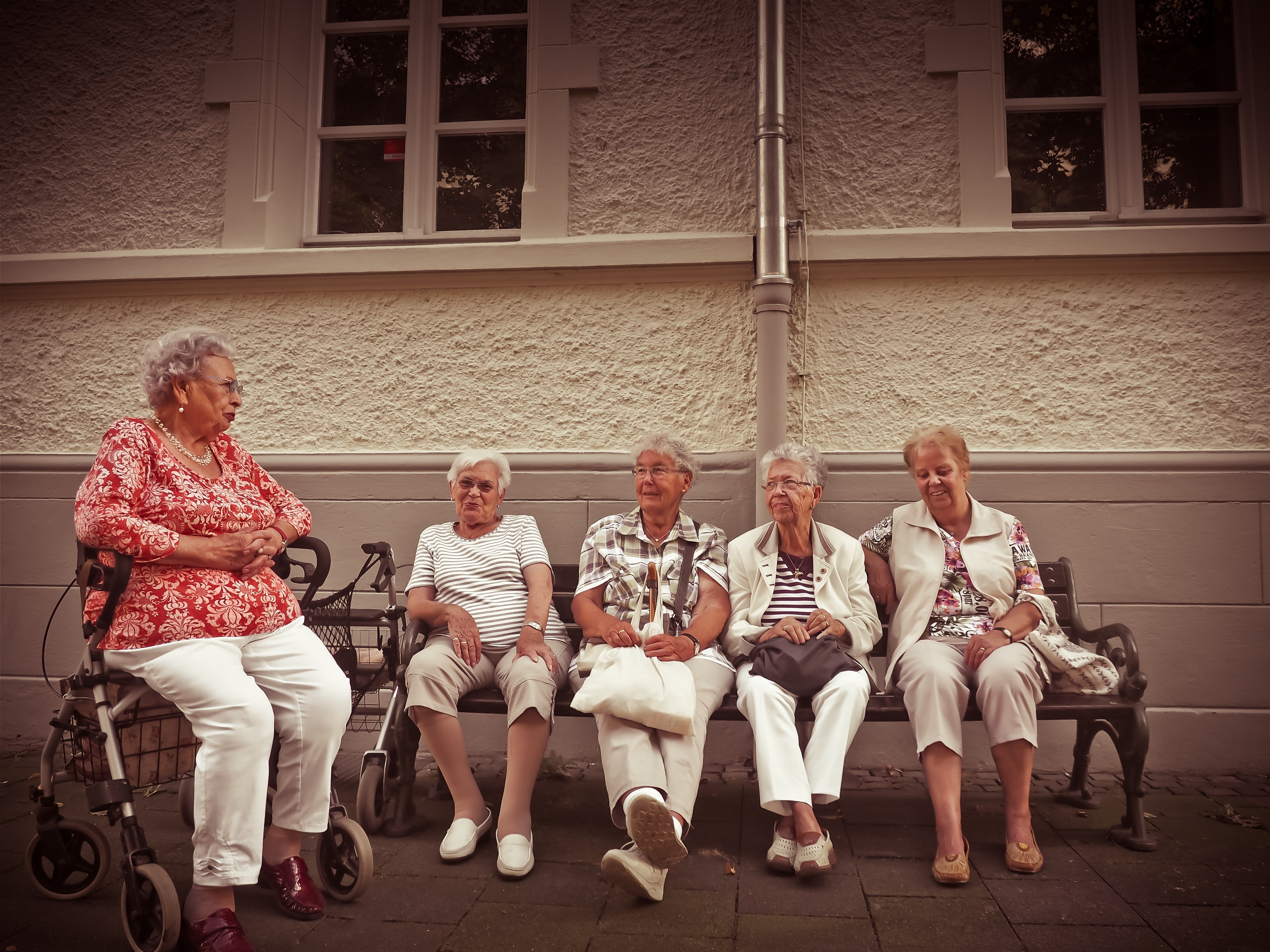 Five elderly women sitting on a park bench outside of a building.