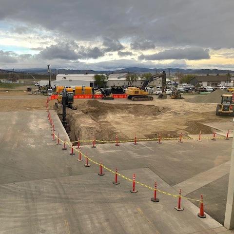 A panoramic view of the liquor warehouse construction site showing a large dirt excavation area with multiple construction vehicles and equipment against a cloudy sky. Semi-trucks and trailers are parked in the distance.